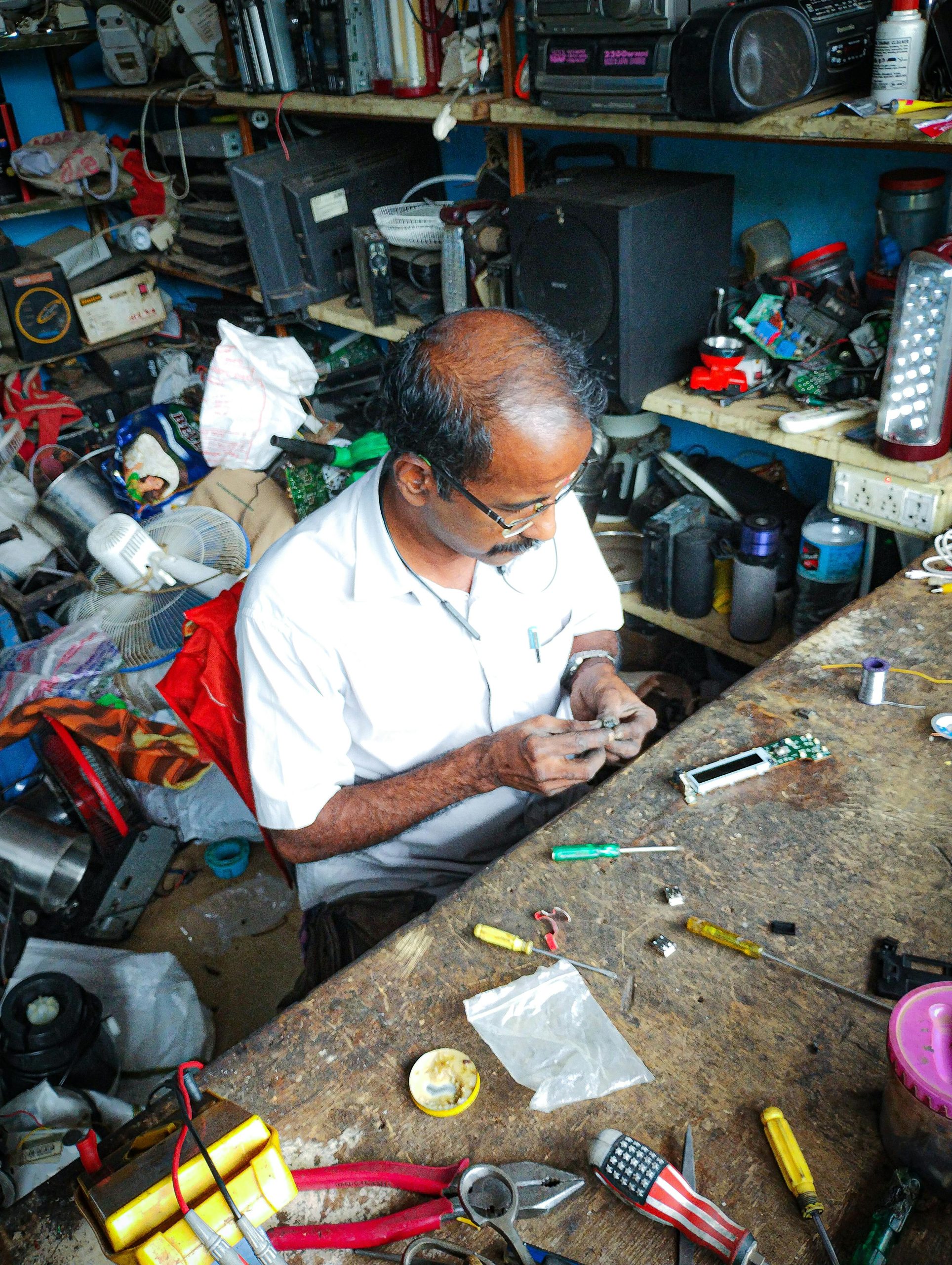 A man repairs electronics in a cluttered workshop, focusing on intricate work.